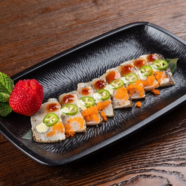 Seared tuna sashimi with sauces, roe, jalapeños, and a carved strawberry on a black plate.