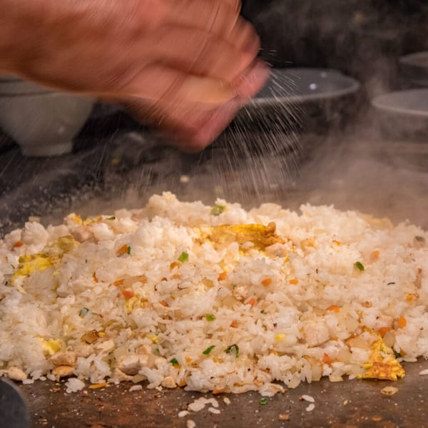 A chef's hand sprinkles seasoning over a mound of Sogo Hibachi fried rice on a grill.