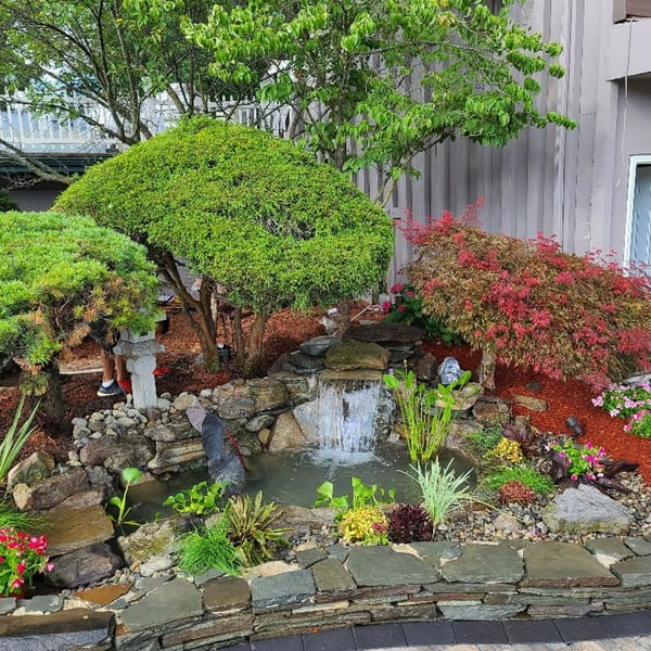 A Japanese garden with a small waterfall, pond, lush green and red foliage, and stone landscaping.