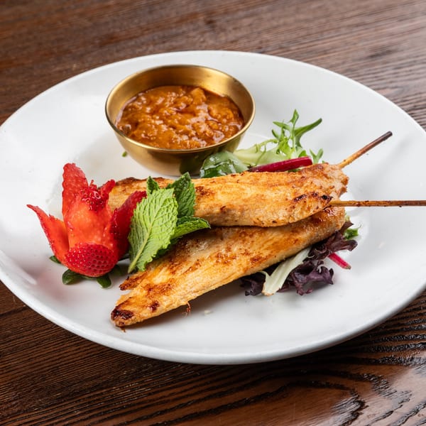 Two grilled chicken skewers with a side of peanut sauce, garnished with mint, greens, and a strawberry.